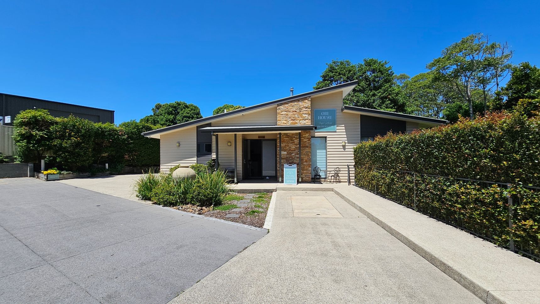 A modern, single-storey building with a slanted roof and a mix of stone and light-coloured siding. The entrance features a small porch with a sign and two chairs. A paved driveway leads up to the building, with neatly trimmed hedges on the right and a small garden area with plants on the left. Trees and greenery are visible in the background.