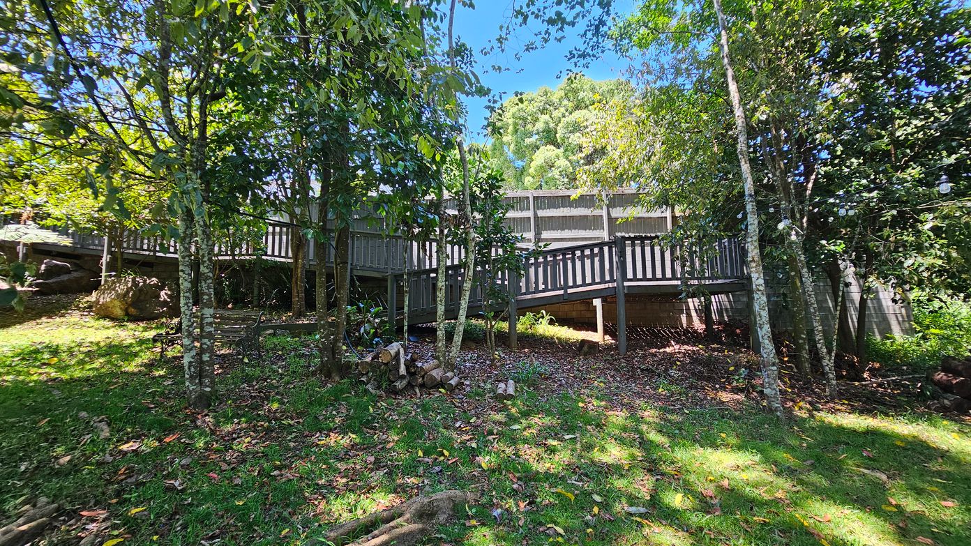 A wooden walkway elevated on stilts runs horizontally across the middle of a lush, green garden. The foreground is covered with grass and scattered leaves, with a few logs piled near the centre. Tall trees with dense foliage surround the area, casting dappled shadows on the ground. In the background, a wooden fence is partially visible through the trees. The scene is brightly lit, suggesting a sunny day.
