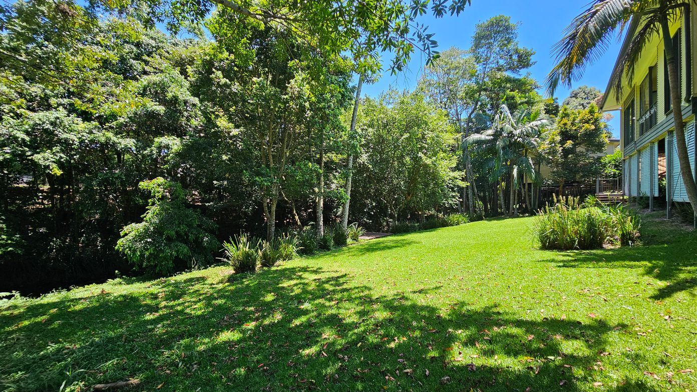 A lush garden with a well-maintained green lawn stretches across the foreground. On the left, dense trees and shrubs create a natural border. To the right, a house with a wooden exterior and a small deck is partially visible, surrounded by tall palm trees. The background is filled with more trees, providing a sense of seclusion and tranquillity. Shadows from the trees dapple the grass, adding texture to the scene.