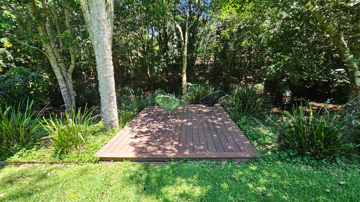 A wooden deck is situated in a lush garden setting. Two chairs, one green and one black, are placed on the deck. Surrounding the deck are tall trees and dense greenery, with various plants and shrubs. The ground is covered with grass, and sunlight filters through the leaves, casting dappled shadows on the deck.