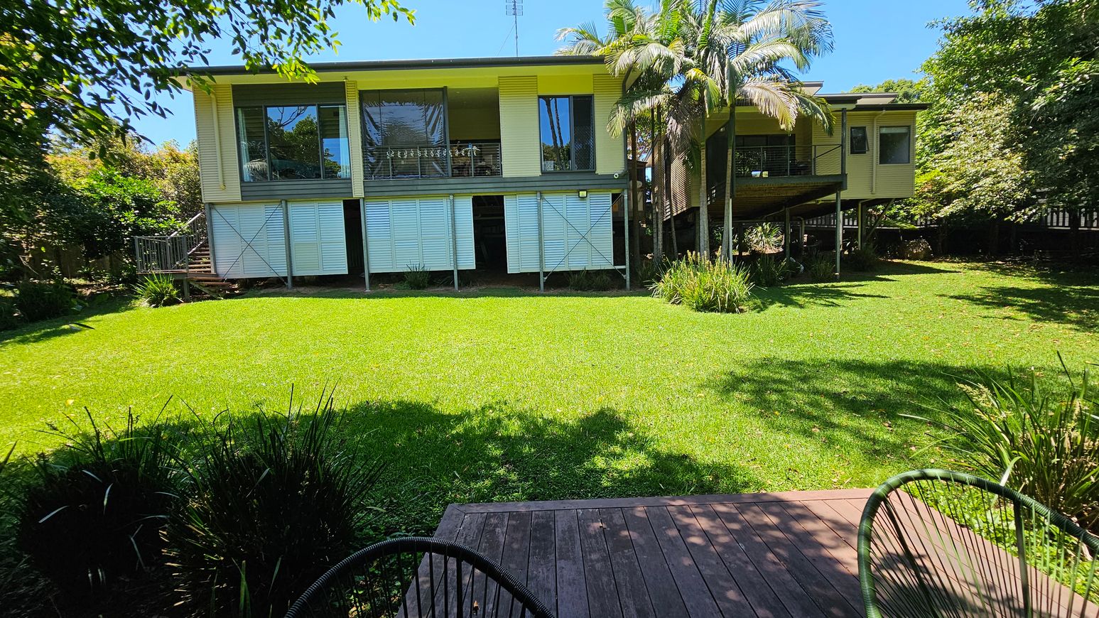 A house elevated on stilts with a wooden exterior and large windows. The foreground features a wooden deck with a green chair. The garden is lush with a well-maintained lawn and various plants. On the right, there are tall palm trees near the house. The background is filled with dense greenery, including trees and shrubs.
