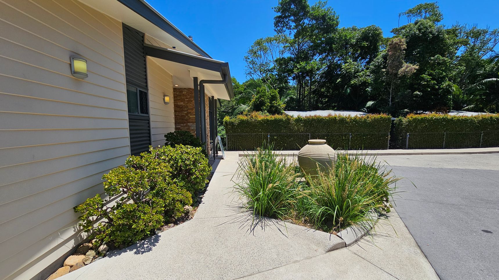 A modern building with light-coloured horizontal siding on the left, featuring a small window and exterior light. Bushes line the base of the wall. In the centre, a concrete path leads to a circular garden bed with tall grasses and a large decorative urn. To the right, a paved driveway curves around the garden bed. In the background, a tall hedge and dense trees provide a natural backdrop.