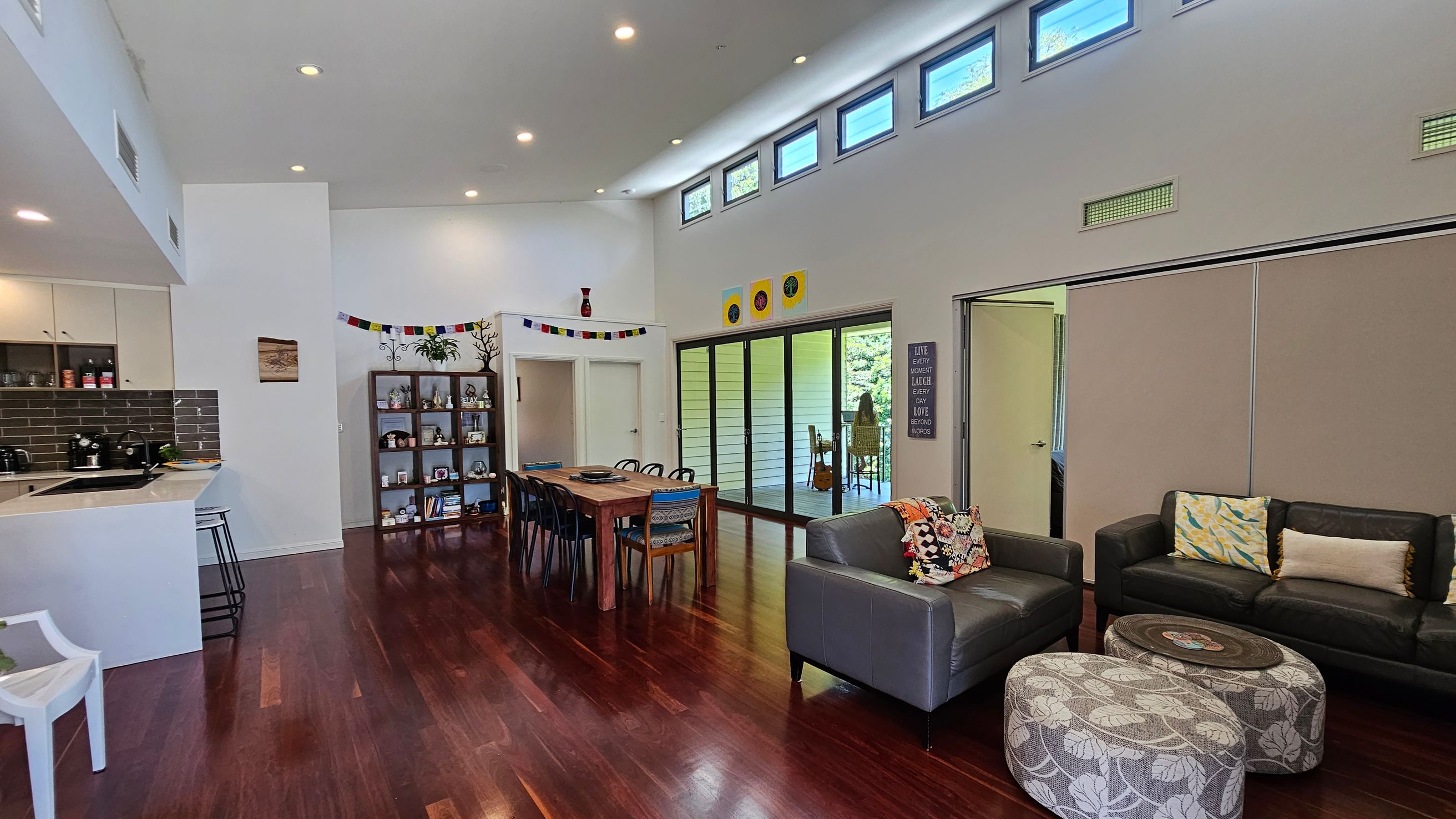 A spacious living area with dark wooden flooring. On the left, there's a kitchen with a white counter and bar stools. In the centre, a wooden dining table with chairs is set near a bookshelf filled with various items. The right side features a grey sofa with colourful cushions and two patterned ottomans. Large sliding glass doors lead to an outdoor area. The ceiling is high with small windows near the top, allowing natural light to enter.
