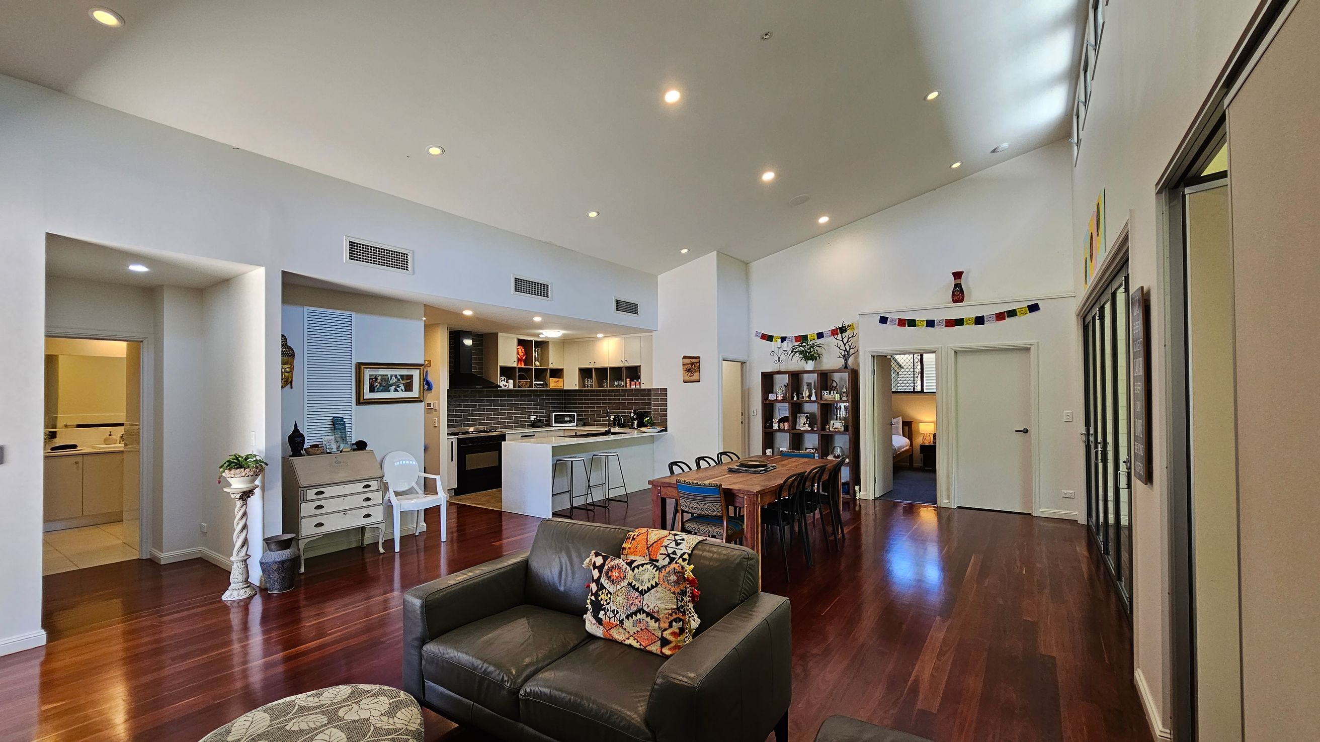 A spacious living area with wooden flooring and a high ceiling. On the left, there's a small bathroom entrance and a decorative plant stand. The centre features a kitchen with a bar counter and stools, and a dining table with chairs. On the right, a black leather sofa with a patterned cushion faces the room. In the background, a bookshelf and a doorway leading to a bedroom are visible. The walls are adorned with colourful flags and artwork.