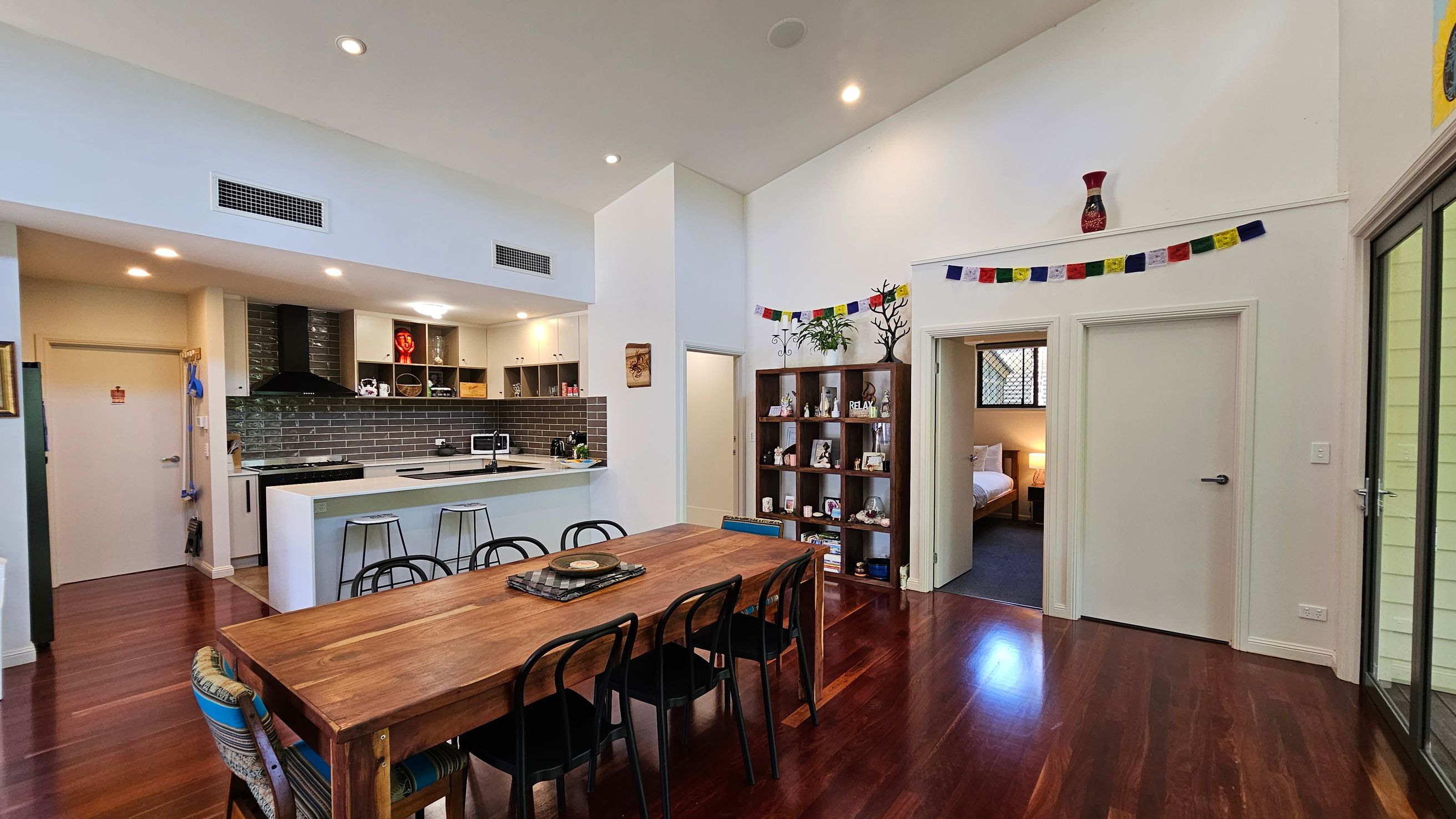 Open-plan living area with a wooden dining table and black chairs in the foreground. To the left, a modern kitchen features a white island with stools, dark tiled backsplash, and open shelving. In the background, a bookshelf with decorative items and a string of colourful flags above. A doorway leads to a bedroom with a visible bed. The floor is polished wood, and the ceiling is high with recessed lighting. Sliding glass doors are on the right.
