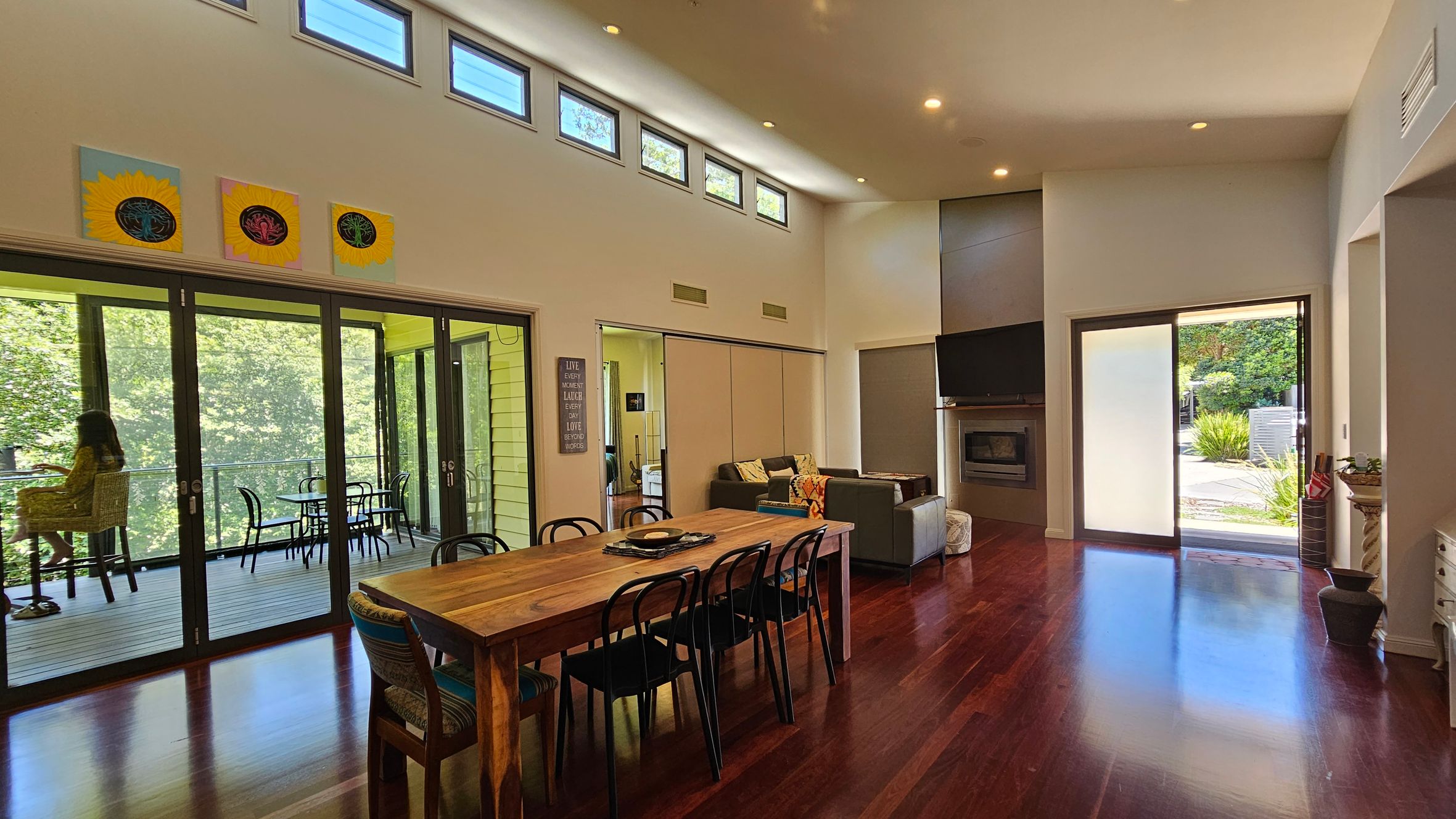 A spacious living room with wooden flooring and a high ceiling. On the left, large glass doors open to a balcony with a person sitting and a table with chairs. Above the doors, there are small rectangular windows and three sunflower-themed artworks. In the centre, a wooden dining table with black chairs. On the right, a seating area with a sofa, a wall-mounted TV above a fireplace, and a sliding glass door leading outside.