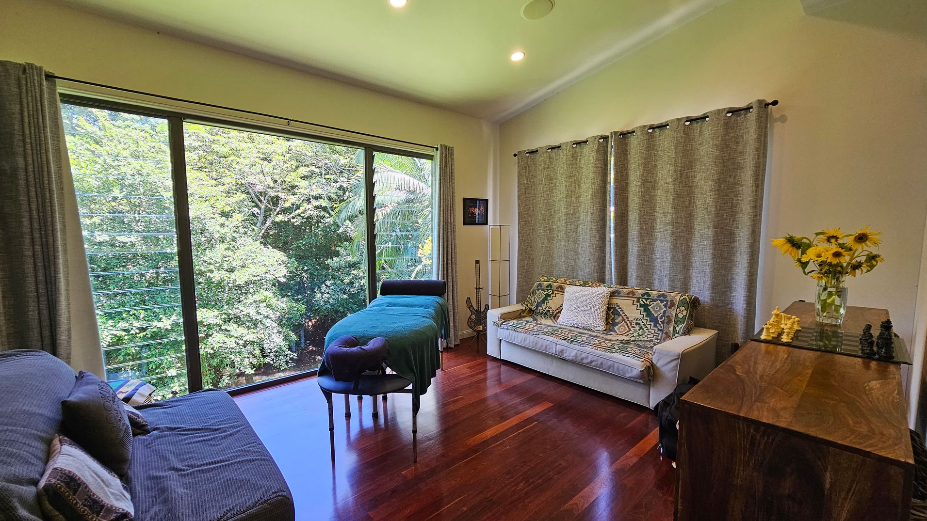 A room with wooden flooring and a large window on the left, offering a view of lush green trees. In front of the window is a massage table with a teal cover. To the right, a white sofa with patterned cushions is placed against a wall with grey curtains. A wooden cabinet on the far right holds a vase of sunflowers and a chessboard. The ceiling is light-coloured with recessed lighting.
