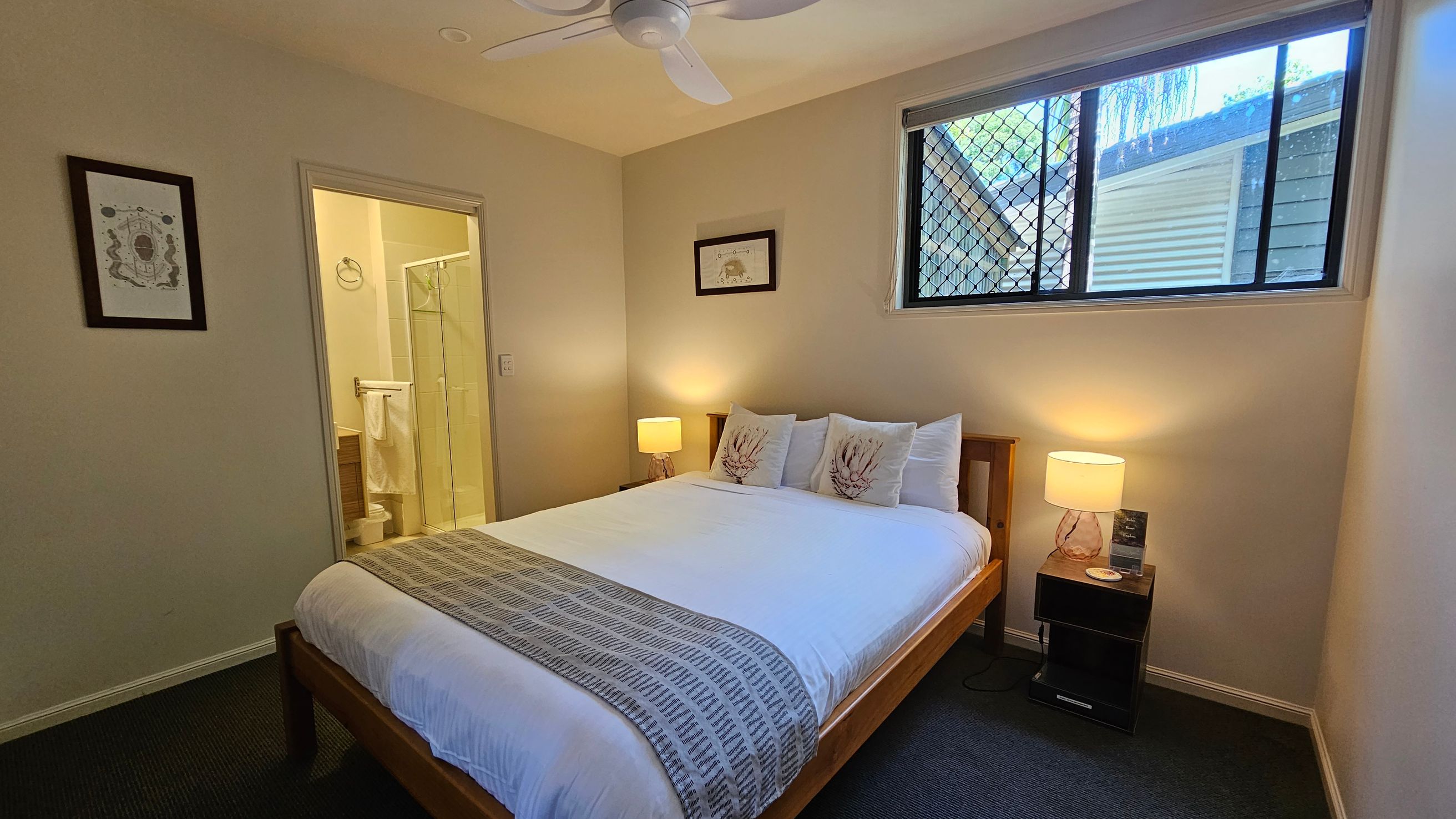 A bedroom with a large bed in the centre, featuring white bedding and decorative pillows. On either side of the bed are bedside tables with lamps. A window with a mesh screen is on the right wall. To the left, there's a doorway leading to a bathroom with a visible shower and towel rack. The walls are light-coloured, and there's a ceiling fan above. A framed picture hangs on the left wall. The floor is carpeted in a dark shade.