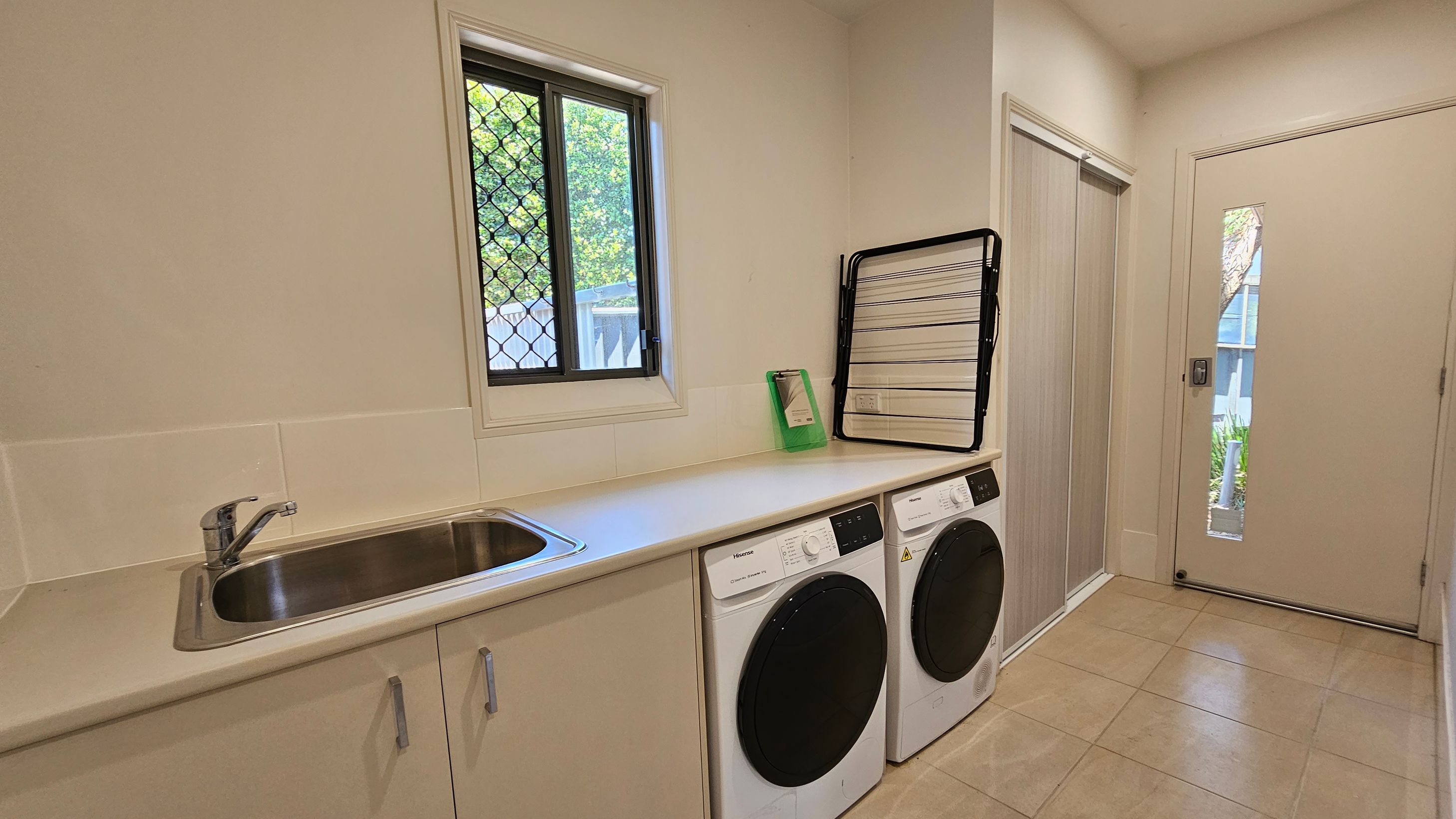 A laundry room with a tiled floor and light-coloured walls. On the left, there's a countertop with a stainless steel sink and cabinets below. A window with a security screen is above the counter. Below the counter are a washing machine and a dryer. On the right, a door with a narrow window leads outside. A folded drying rack and a green clipboard are on the counter. The ceiling is plain and light-coloured.