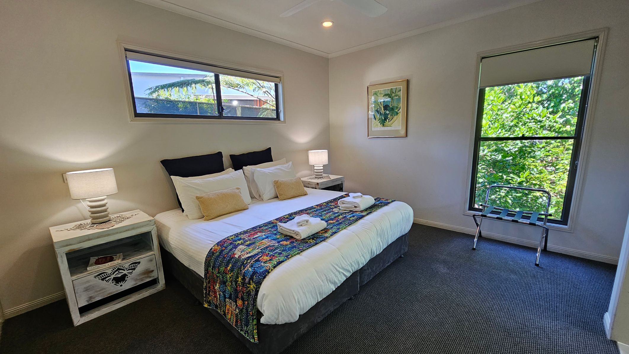 A bedroom with a large bed in the centre, adorned with white linens, two beige cushions, and a colourful throw. Two neatly folded towels are placed on the throw. On either side of the bed are white bedside tables with lamps. A small window above the bed and a larger window on the right let in natural light, with greenery visible outside. A luggage rack is positioned near the larger window. The floor is carpeted in a dark grey tone, and a framed picture hangs on the wall.