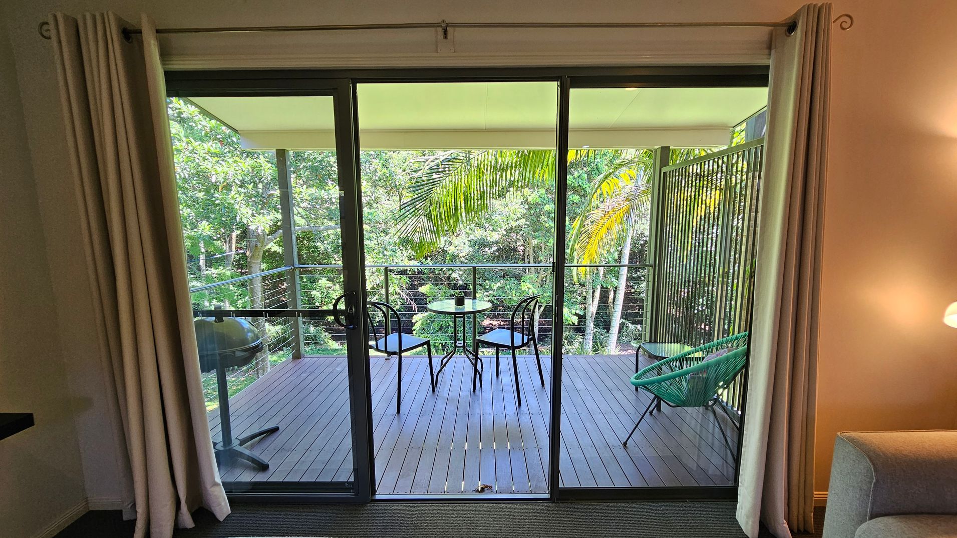 A view through glass sliding doors leading to a wooden deck. On the deck, there's a small round table with two black chairs and a green chair to the right. A barbecue grill is on the left. The background features lush green trees and palm leaves. Inside, beige curtains frame the doors, and a grey sofa is partially visible on the right. A lit wall lamp is above the sofa.