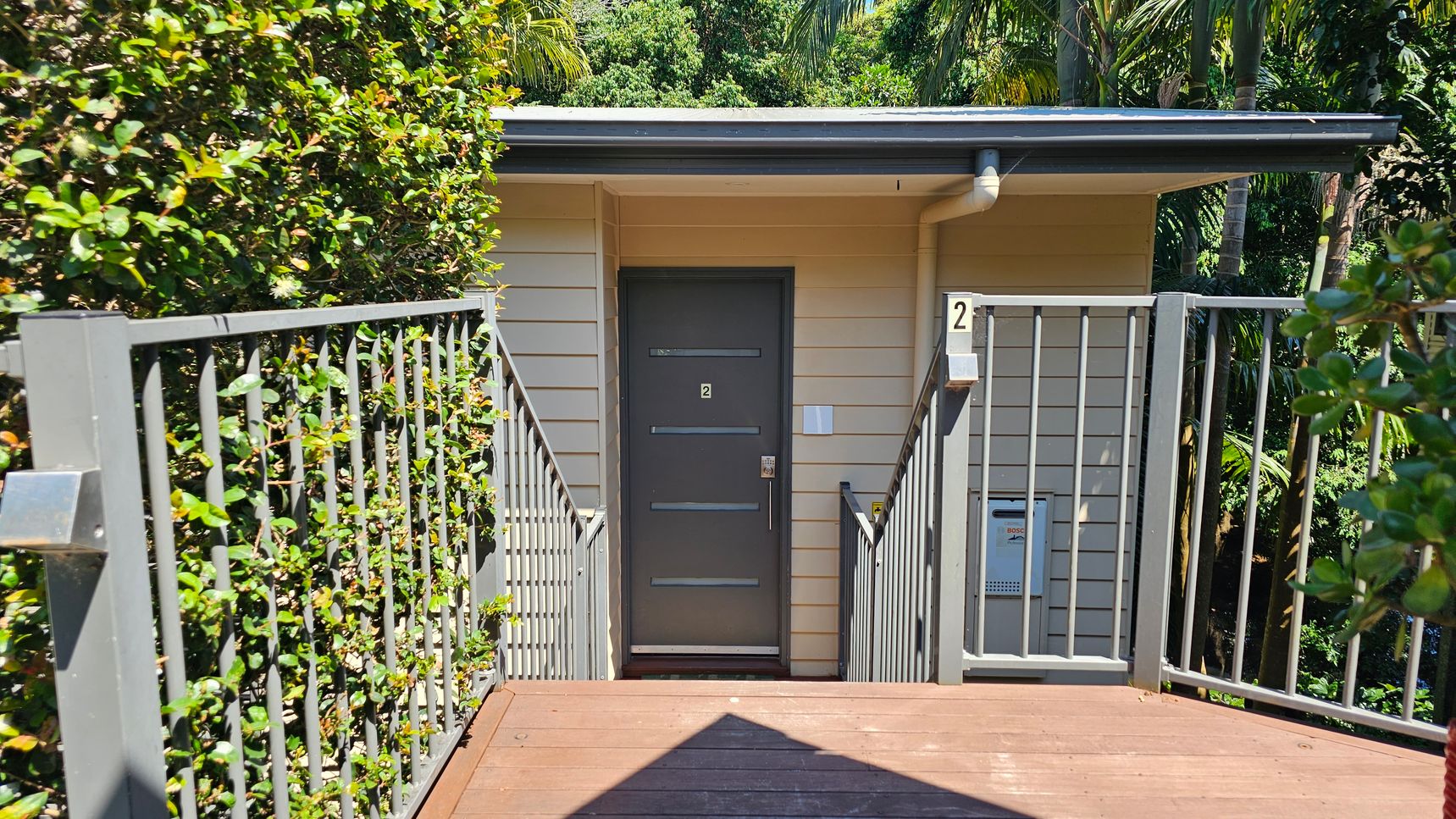 A wooden walkway leads to a grey door with the number "2" on it. The walkway is flanked by metal railings entwined with green foliage. The building is clad in light-coloured horizontal siding. To the right of the door, a small utility box is visible. Lush greenery and trees are in the background, creating a natural setting.