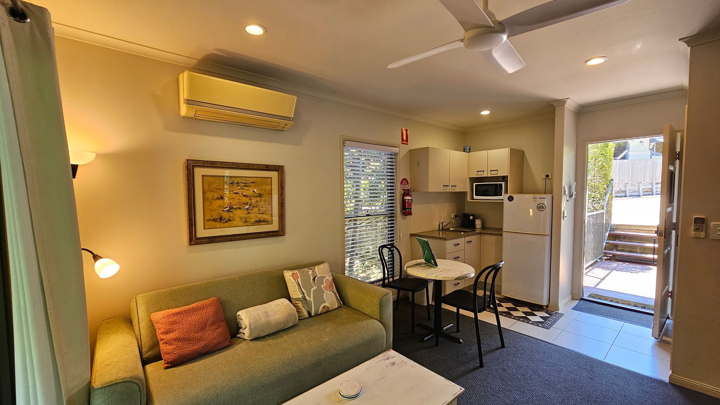 A cosy living area with a green sofa adorned with patterned and plain cushions on the left. Above the sofa is a framed painting and a wall-mounted air conditioner. A small round table with two black chairs is in the centre. The kitchenette on the right features a fridge, microwave, and cabinets. A door opens to a sunlit outdoor area with steps leading up. The floor is a mix of carpet and tiles, and a ceiling fan is visible above.