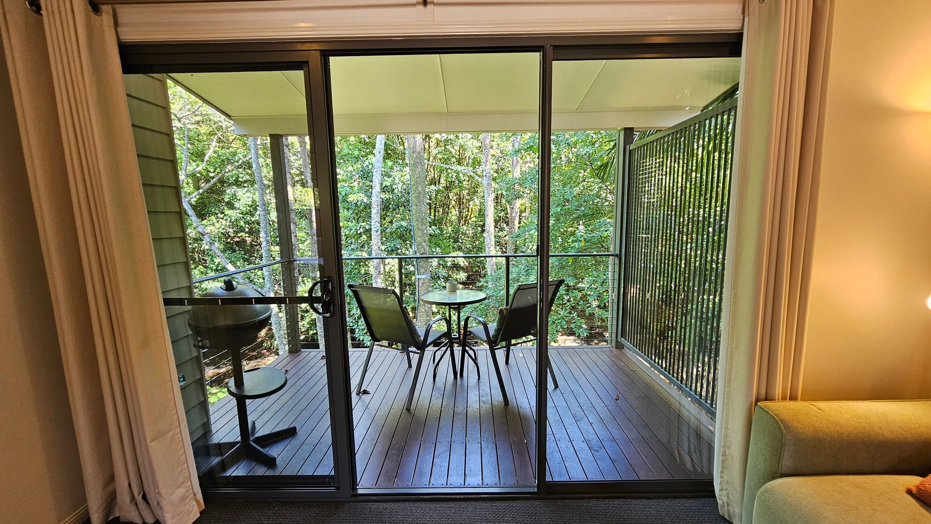Sliding glass doors open to a wooden deck with a small round table and two chairs. A barbecue grill is on the left. The deck is surrounded by lush green trees. Inside, beige curtains frame the doors, and a light green sofa is partially visible on the right. The ceiling is white, and the floor inside is carpeted.