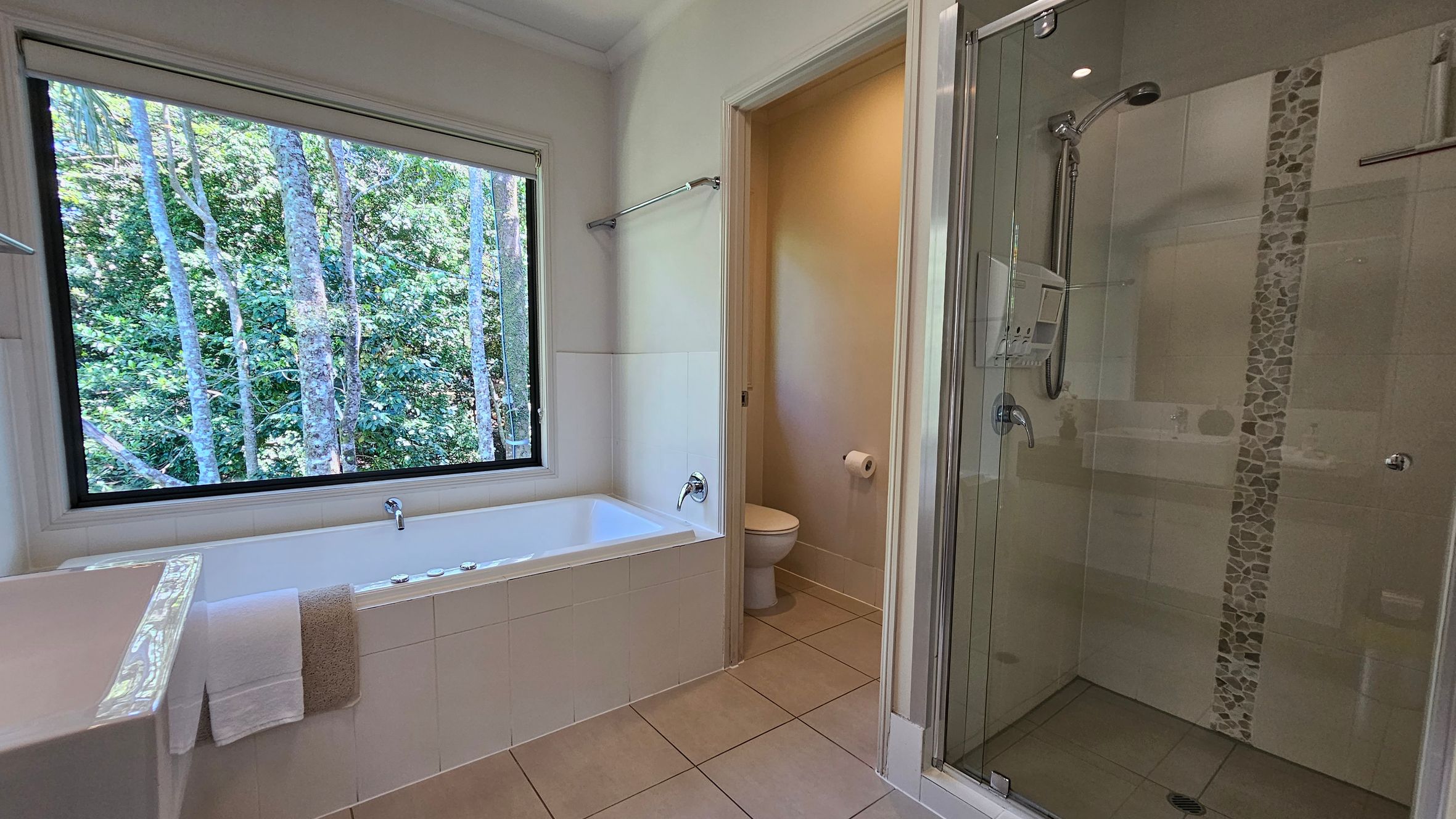 A bathroom with a large window on the left, offering a view of trees outside. Below the window is a white bathtub with a towel draped over the edge. In the centre, there's a doorway leading to a toilet area. On the right, a glass-enclosed shower with a pebble-tiled vertical strip. The floor is tiled in a light colour, and the walls are painted white.