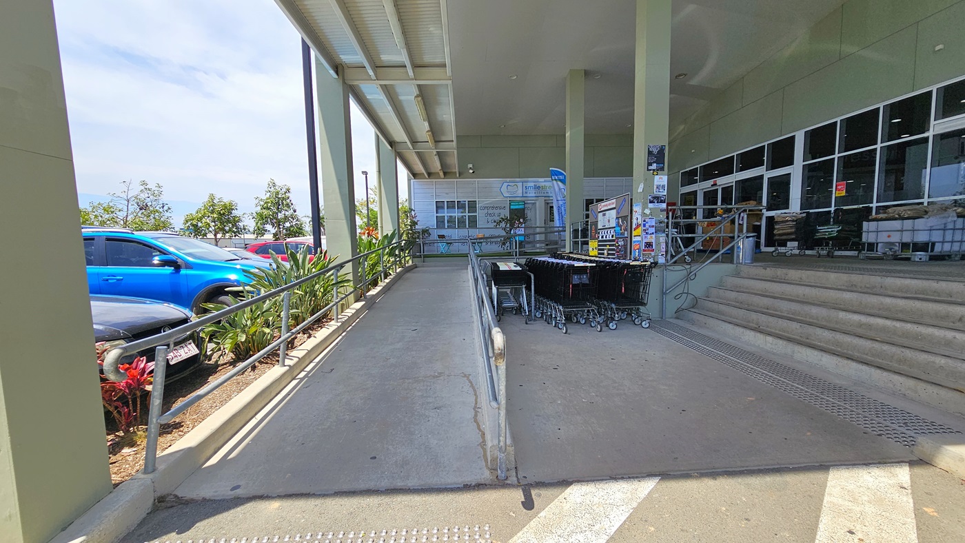 A closer view of the ramped and 5-step staircase entrances both with truncated domes before entering. Safety handrails are placed at both entrances. Adjacent to the shopping trolleys, posters and signage are placed on a nearby wall. Going to the ramped entry leads to where “smile street” is situated.
