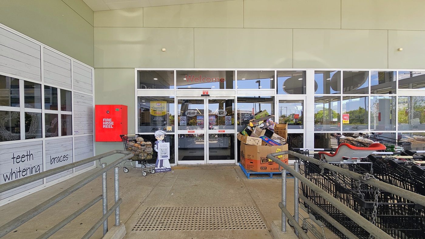 Entrance leading to the inside of Tweed Valley IGA with truncated domes on the floor surface. A Cérge standee is holding a QR code is placed adjacent to a shopping trolley filled with vegetables near the glass doors. A fire hose reel is visibly placed in the left corner. A stack of used boxes sits near the right side of the entrance.