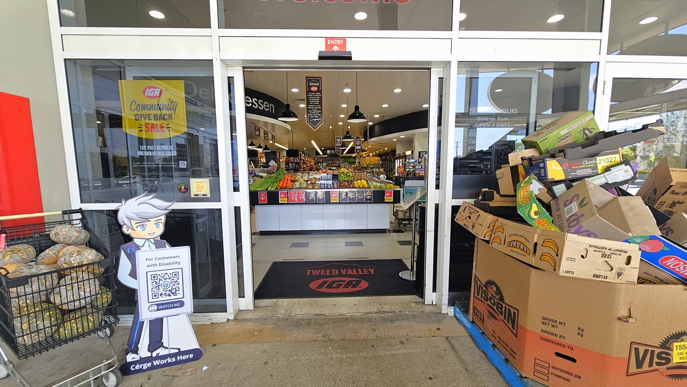 Entrance of the Tweed Valley IGA building. A black doormat with the building’s name and icon is visible. A well-organized display of fruits and vegetables is situated at the entrance upon entering. Fluorescent lights and hanging banners clearly dominate the ceiling of the building.