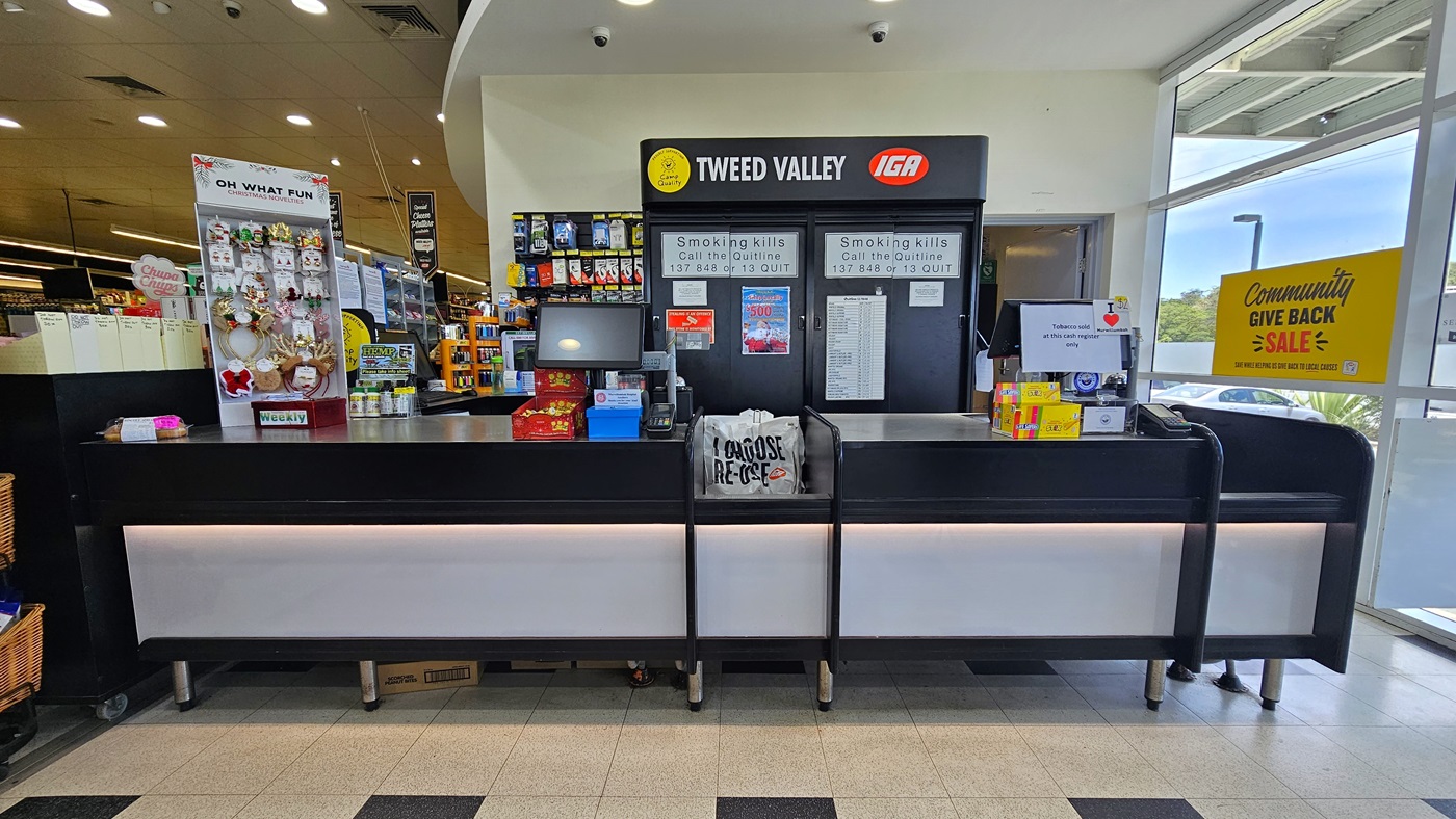Checkout area near the entrance of the building. On the left, a multi-layer rack full of chips and other products is visible beside three piled-up wooden baskets with chips. Two counters are also visible on the left, with three checkout staff members noticeable. On top of the employee's left, there are two functional CCTVs. On the right, there are also three wooden baskets with chips and a fridge full of soft drinks, where several water bottles sit atop.
