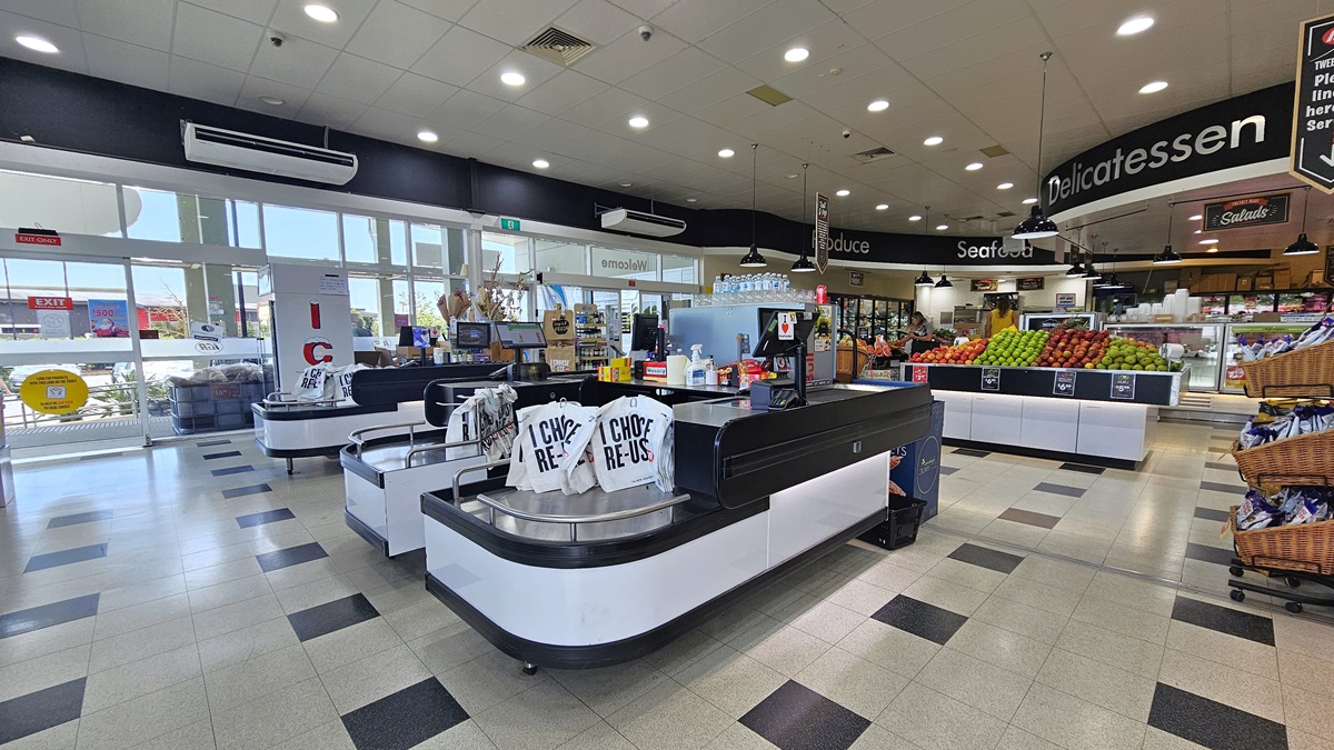 A closer look at the checkout counters. Three checkout counters with the same colors of black and white have reusable bags sitting on top of each of them. Two big air conditioners are visible on top of a wall, near the entrance. On top of the three checkout counters, various products are apparent. Several CCTVs are prominently placed on top of the ceiling.