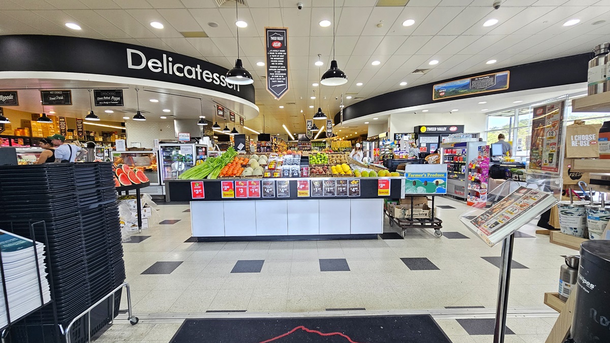 Closer view of the Tweed Valley’s main entrance with a well-organized display of fruits and vegetables. Shopping black baskets are piled up on the left. On the right hand, a stand holding pamphlets and flyers are apparent. Going further inside, the Meat, Produce, and Seafood sections are visible. Bright fluorescent lights illuminate the entire area.