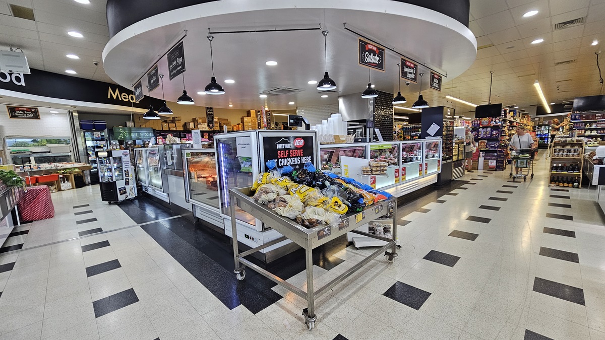 A closer look at the Delicatessen section. “Self-serve chickens” are visibly placed in the center of the Delicatessen near a steel cart loaded with products. Consumers are apparent on the right side corner adjacent to the three-layer shelves. Fluorescent lights and banners overcome the ceiling of the Delicatessen section. Striking black tiles conquer both sides of the area as well.