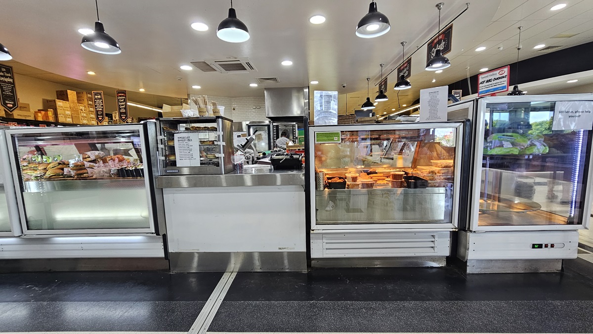 A closer look at the front view of the Delicatessen. Rows of fridges filled with burgers, cooked chicken, and other delicacies are prominent. Names of the delicacies are evidently placed above the fridges on the right. An employee is evident at the far end. Used and unused boxes are noticeable above the shelves on the far left.