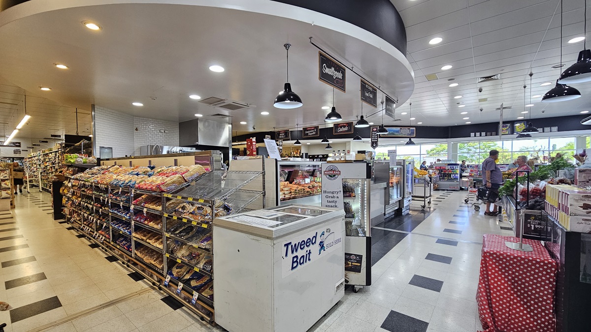 Side view of the Delicatessen section. A freezer is prominently placed adjacent to a row of multi-layer racks filled with bread. A fridge full of beverages is also apparent near the freezer, directly facing a table covered in a cloth with a mixture of red and white dots. Consumers are noticeable to the far right end.