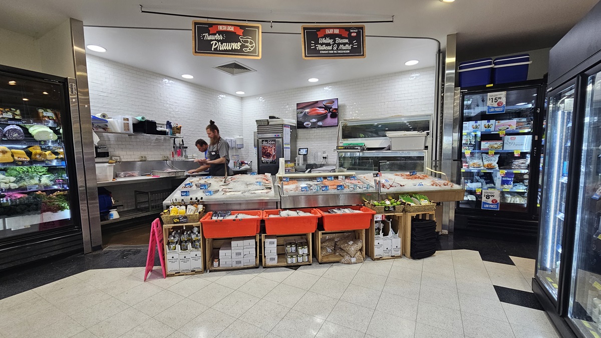 Seafood and meat section. Two banners are noticeably hanging on a black steel rod above the ceiling. Shop assistants are apparent, as well as a wall decor inside the seafood and meat section. Frozen prawns and fish are visibly displayed and underneath are beverages and other products. On the right, two fridges are prominent.