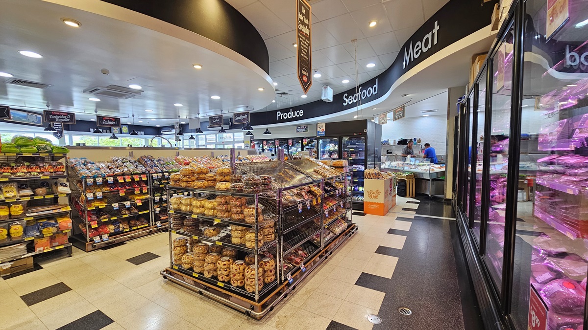 Grocery aisle near the seafood and meat section. Huge labels of product, seafood, and meat sections are visible on top of the ceiling, on a black-colored wall. Delicacies are prominent in the center, beside the racks of bread. On the right, rows of fridges with delicacies are visible. On the far end, on the right side, two shop assistants are apparent inside the seafood and meat section.