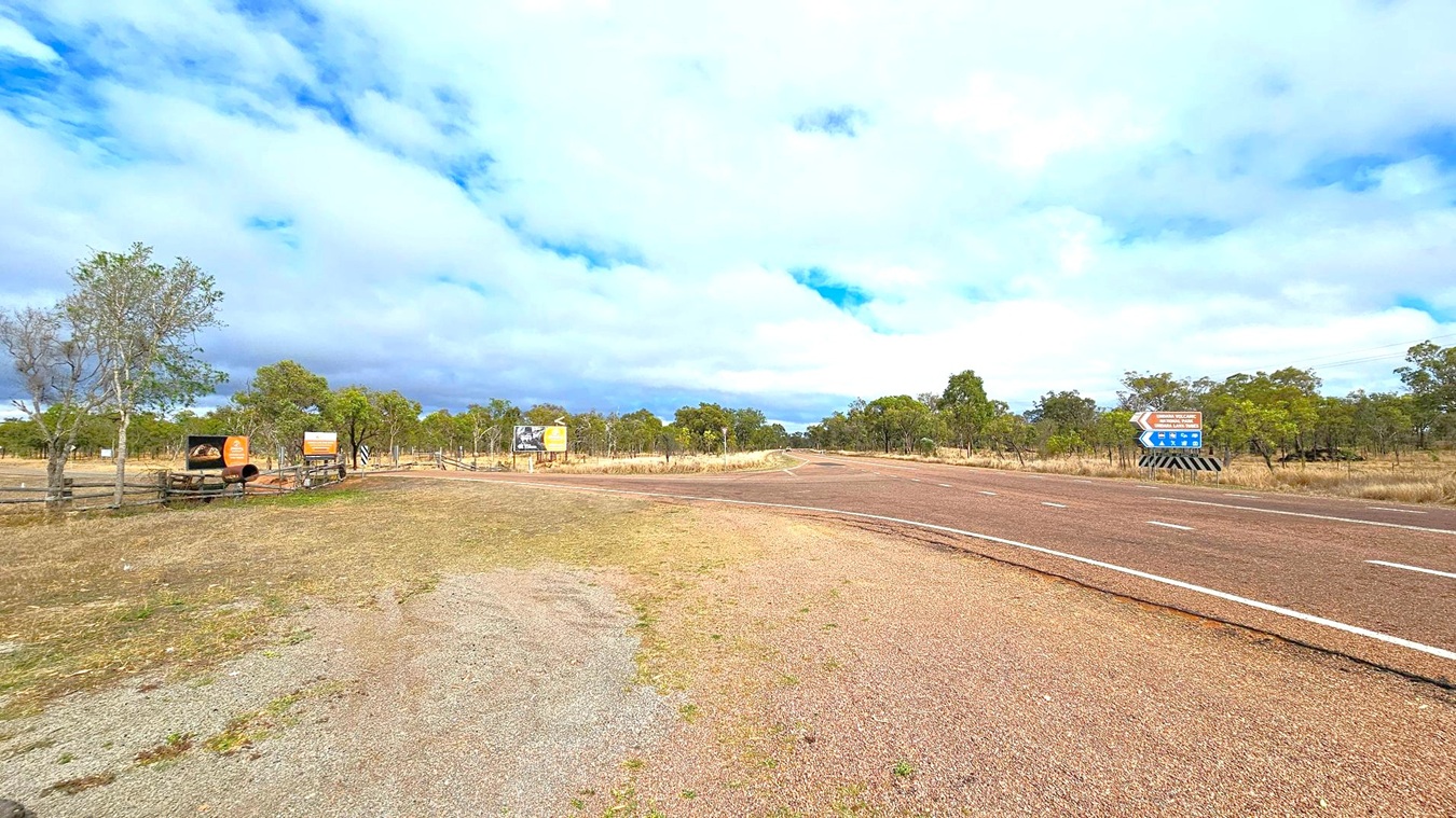 A rural road with a reddish-brown surface curves to the right. On the left, there are several signs, including one with an image of a cow and another with text. Sparse trees and grassland extend into the background. On the right, a blue directional sign with white text and arrows is visible. The sky is mostly cloudy with patches of blue. The ground is a mix of grass and gravel.