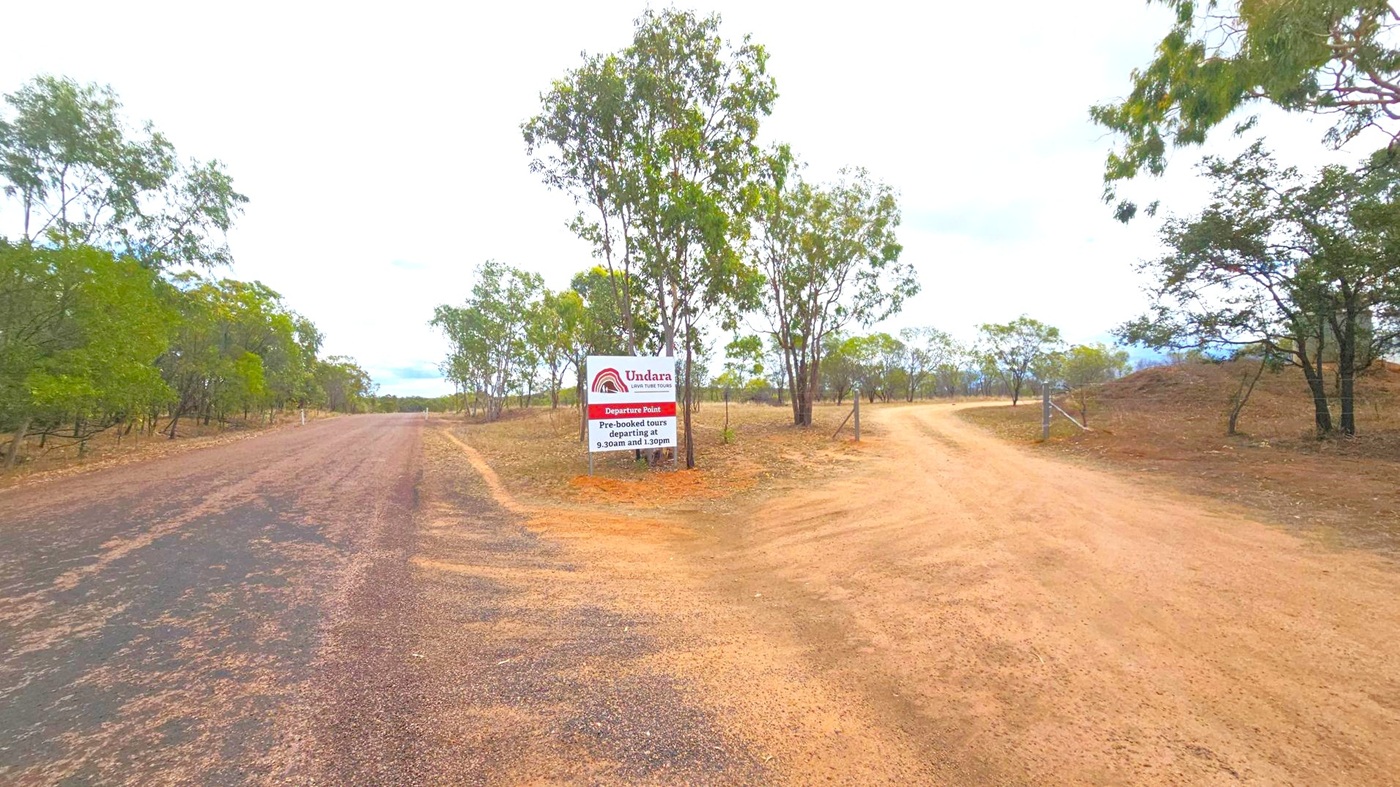 A dirt road on the right curves into the distance, bordered by sparse trees and dry grass. On the left, a wider, darker gravel road runs parallel, also lined with trees. In the centre, a sign reads "Undara Lava Tube Tours" with tour times listed. The sky is overcast, and the landscape is flat with scattered vegetation.