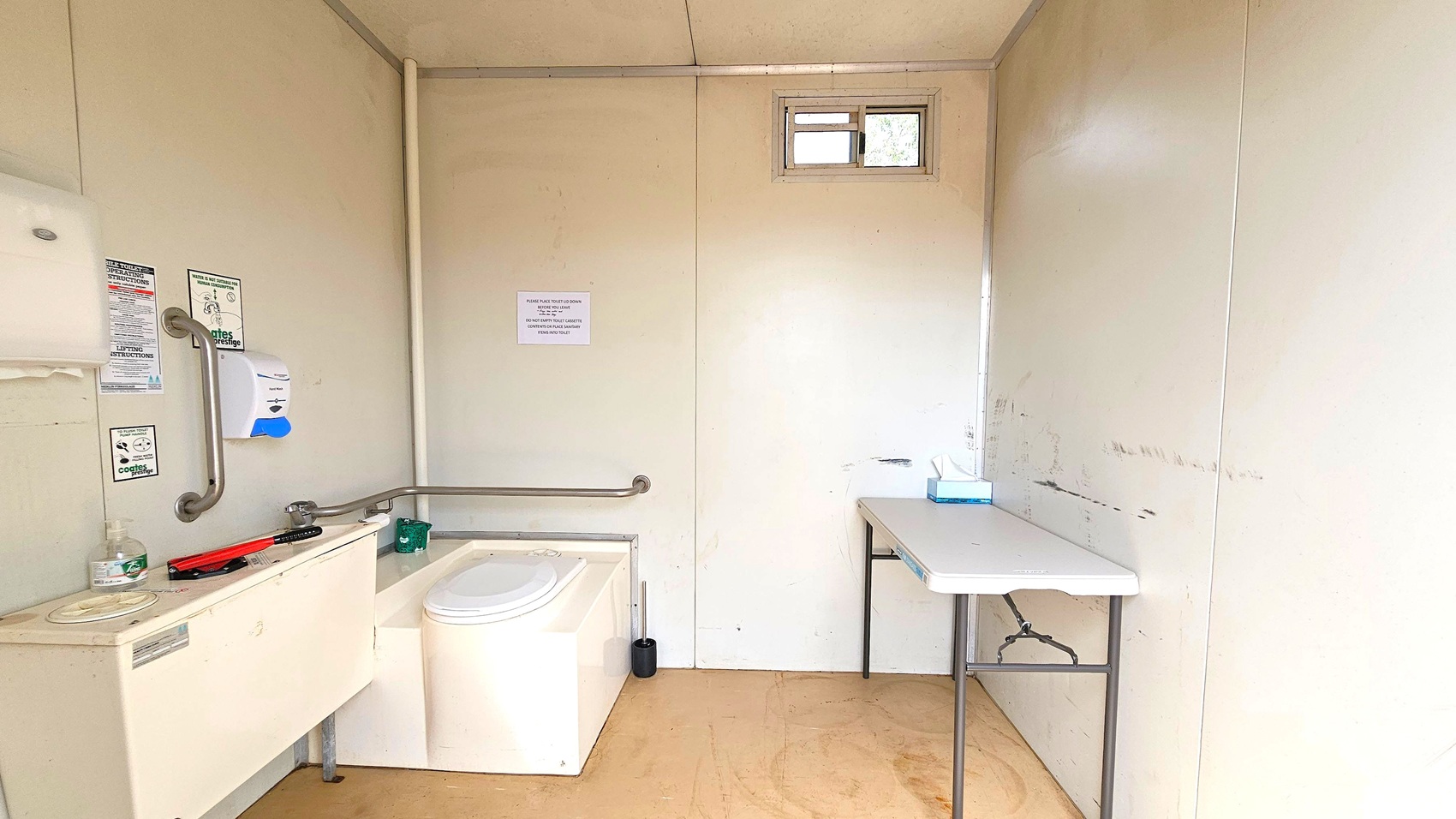 A small, simple bathroom with light-coloured walls and a wooden floor. On the left, there is a white composting toilet with a handrail and a small sink area with a soap dispenser and hand sanitiser. Various instructional signs are posted on the wall. On the right, a folding table holds a box of tissues. A small window near the ceiling allows natural light to enter. The ceiling is plain and matches the walls.