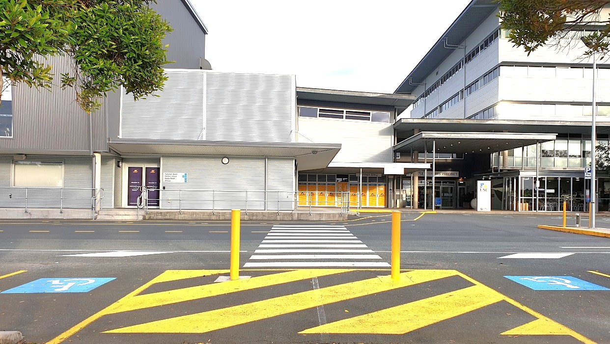 A low-rise building with a grey metal exterior. The left side there are two purple-colored doors with descriptions on them, overlooking a zebra crossing. In the centre are yellow-patterned glass walls and a welcoming inscription on the door. The main entry is a shaded walkway, sign boards, and the USC inscription above a large glass door. Adjacent to it is a see-through glass seating area with signs at the front.