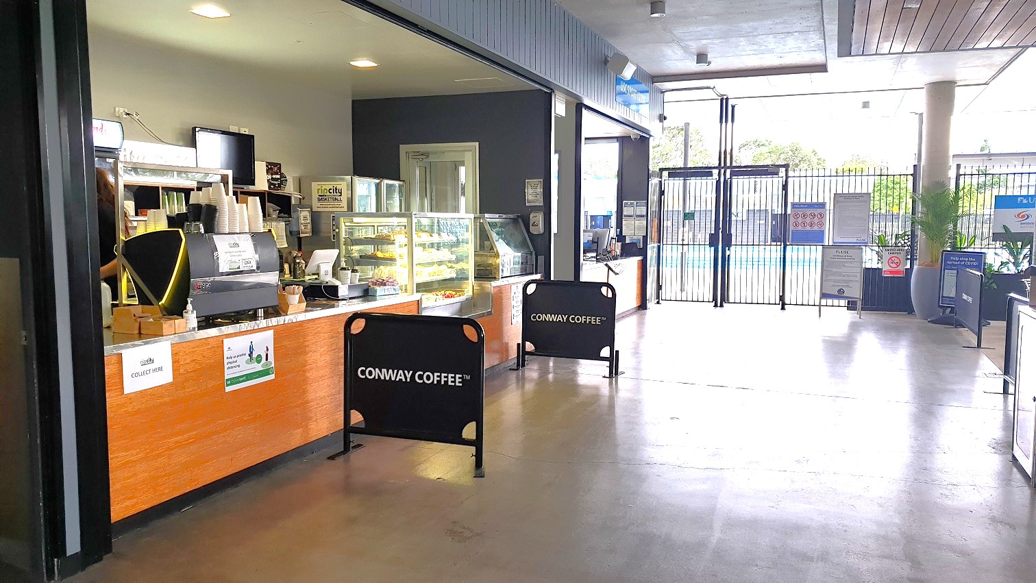 To the left of the see-through fence leading to the pool is a retail area with a café counter and another cubicle with infographics on the wall.  The café has a TV on the wall, a reception counter stocked with snacks in a glass cabinet, an ice cream machine and cups, a desktop, and a few boxes. There are Conway Coffee signage at near the counter for customers to queue alongside. A turnstile gate is to the right, with a large palm in a grey pot beside the pillar with signage on the wall.