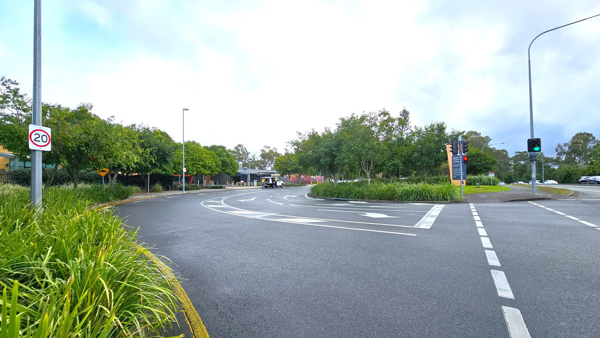 A road with a speed limit sign of 20 on the left, bordered by lush green grass and trees. The road curves to the right, with a white vehicle visible in the distance. On the right, there are traffic lights showing green, and a large sign with text. The road markings indicate lanes and directions. The background is filled with more trees and a cloudy sky.