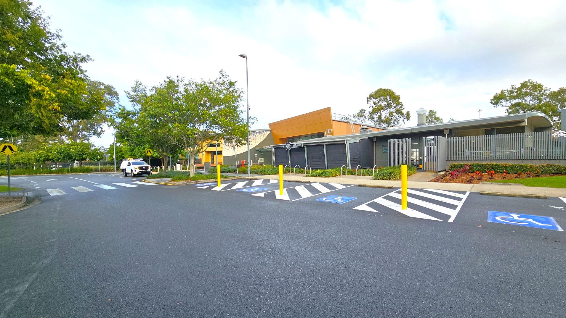 A car park with designated disabled parking spaces marked in blue and white is in the foreground. Yellow bollards separate the spaces. On the right, a modern building with grey and orange panels is visible, surrounded by landscaped gardens. In the background, a white vehicle is parked near a pedestrian crossing. Trees with green foliage line the left side, and a lamppost stands near the centre. The road surface is smooth and dark grey.