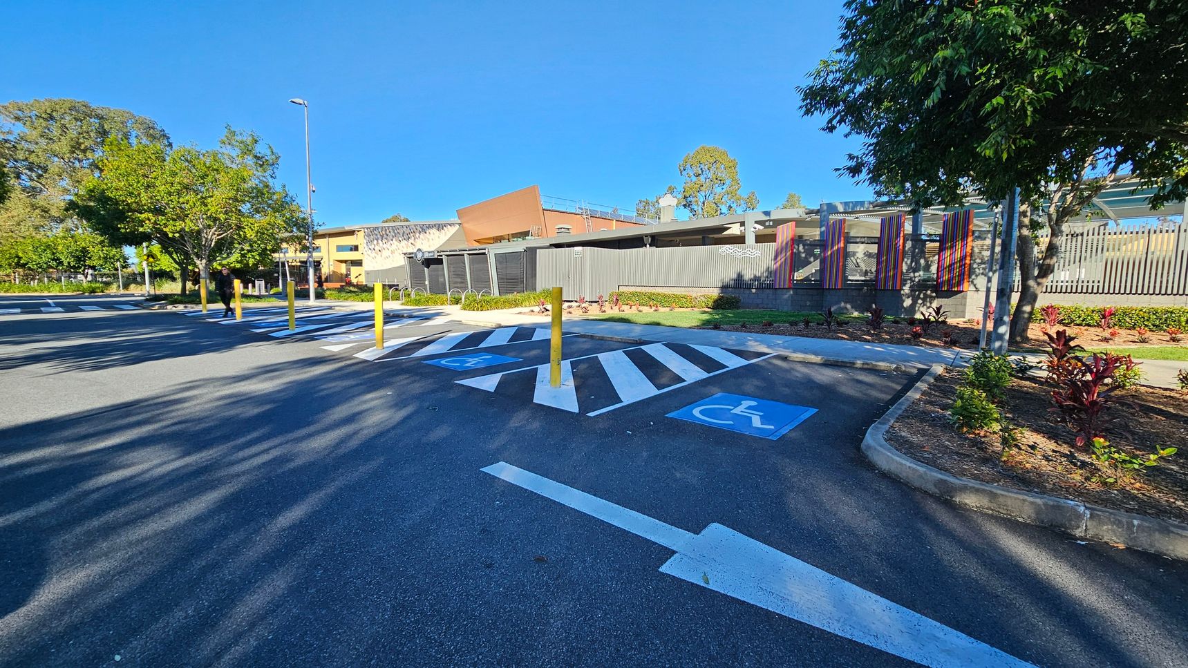 A car park with designated disabled parking spaces marked in blue on the asphalt. White arrows and lines indicate traffic flow and parking boundaries. Yellow bollards are positioned near the spaces. On the right, there are landscaped areas with small plants and trees. In the background, a modern building with colourful vertical panels and a fence is visible. Trees and a lamppost are on the left side. Shadows from the trees stretch across the ground.