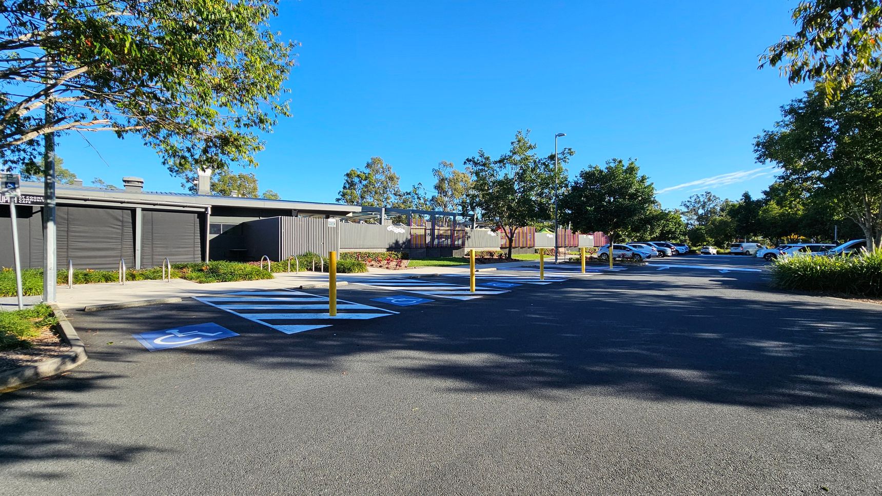 A car park with designated disabled parking spaces marked in blue and white on the asphalt. Yellow bollards separate the spaces. On the left, a modern building with dark panels and a small garden area. Trees with green foliage are scattered throughout, providing shade. In the background, more parked cars and additional trees. The ground is paved, and the area is well-maintained.