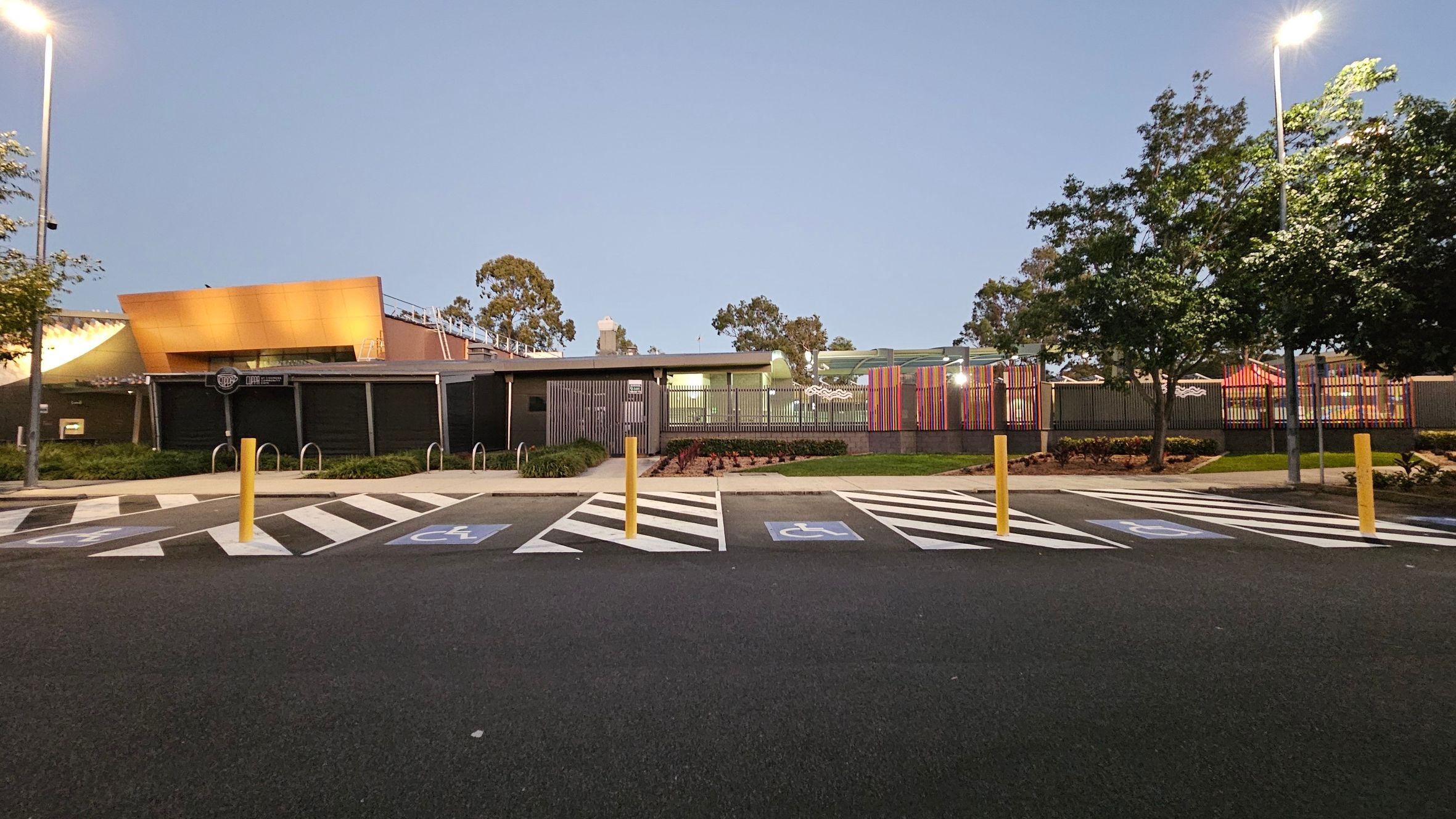 A parking area with marked disabled spaces in the foreground, featuring blue and white symbols. Yellow bollards separate the spaces. In the background, a modern building with a golden-brown facade and a section with colourful vertical slats. Trees are on both sides, and streetlights illuminate the scene. The ground is asphalt, and the area is well-maintained with some greenery.