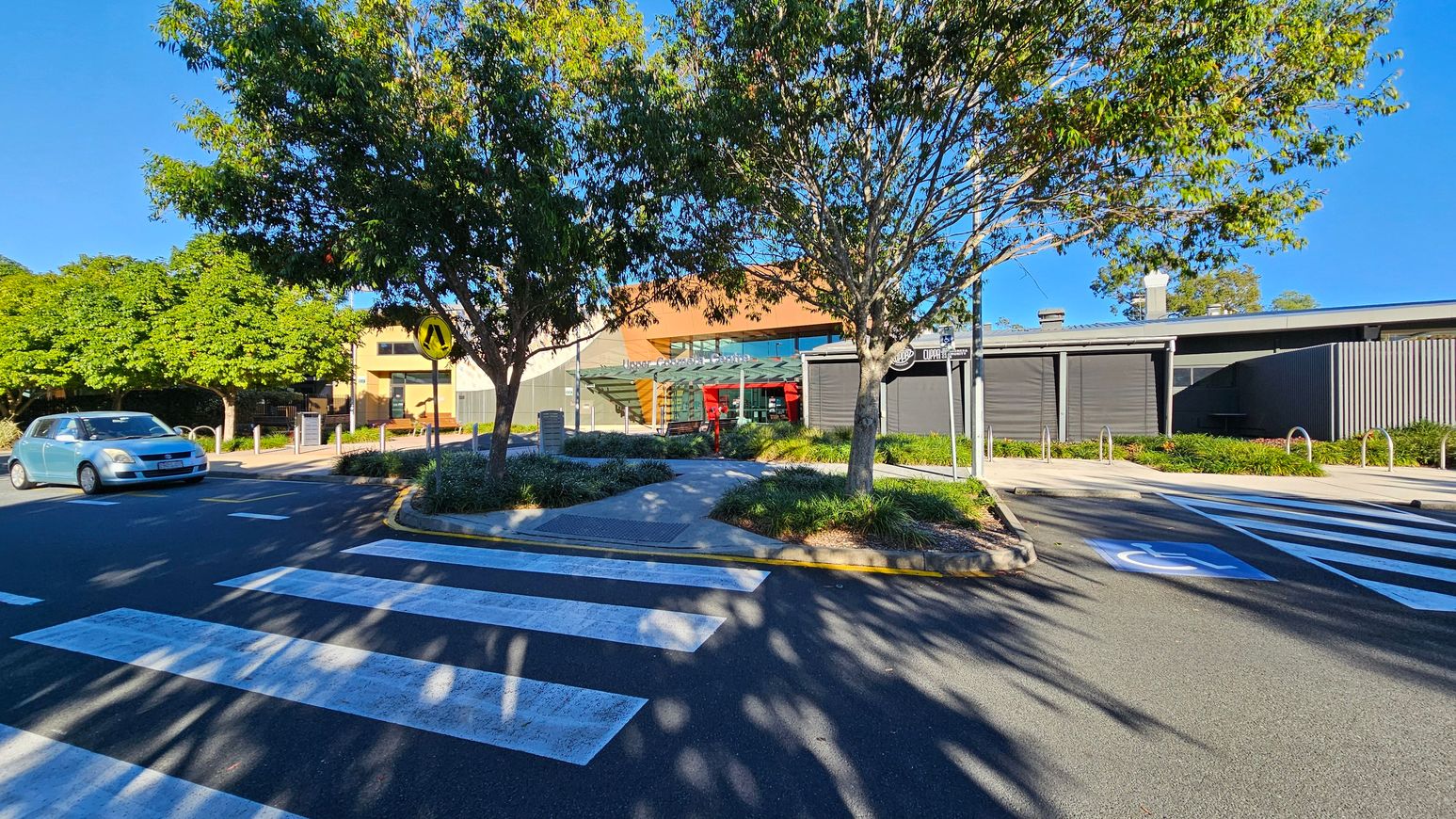 A zebra crossing leads to a pathway flanked by two trees with green foliage. A light blue car is on the left, approaching the crossing. In the background, a modern building with a glass entrance and red accents is visible. The area is surrounded by well-maintained greenery and a signpost is near the pathway. The road has a marked disabled parking space. Shadows of the trees are cast on the road.