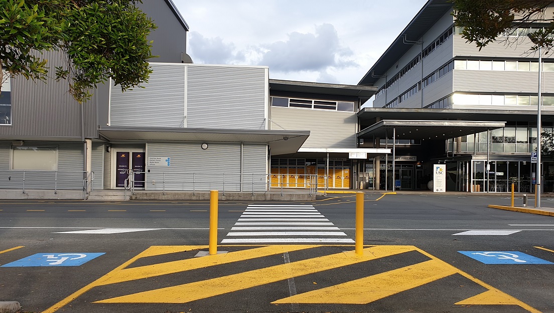 A large, industrial style building with a grey metal façade. On the left side are two purple-colored doors with descriptions on them, overlooking a walkway with pedestrian markings. In the center are yellow-patterned glass walls and a welcoming inscription on the door. The main entry is a well-lit grey façade with a roll-up banner and the USC inscription above a large glass door. Adjacent to it is a see-through glass seating area with signs in front.