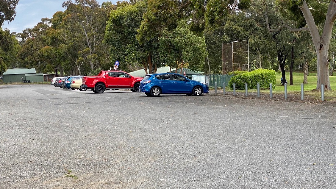 A wide parking space surrounded by trees and marked by short bollards overlooks the entrance to the golf course and the club building to the left. Green-painted metal fence and a golf course fence are seen.  At the left end are two green structures.