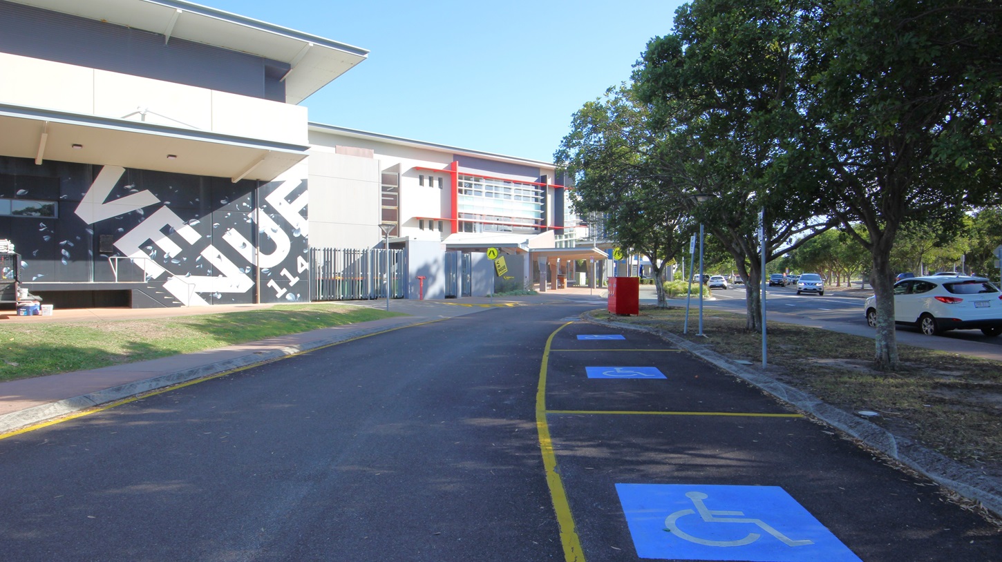 On the left, there is a building with a large sign reading "VENUE" in bold letters, featuring a slanted roof and a grassy area in front. On the right, there are several trees and parked cars along a road. In the middle, a building with red accents and multiple windows extends towards the background. The foreground features a paved road with yellow lines and blue disabled parking spaces marked with wheelchair symbols.