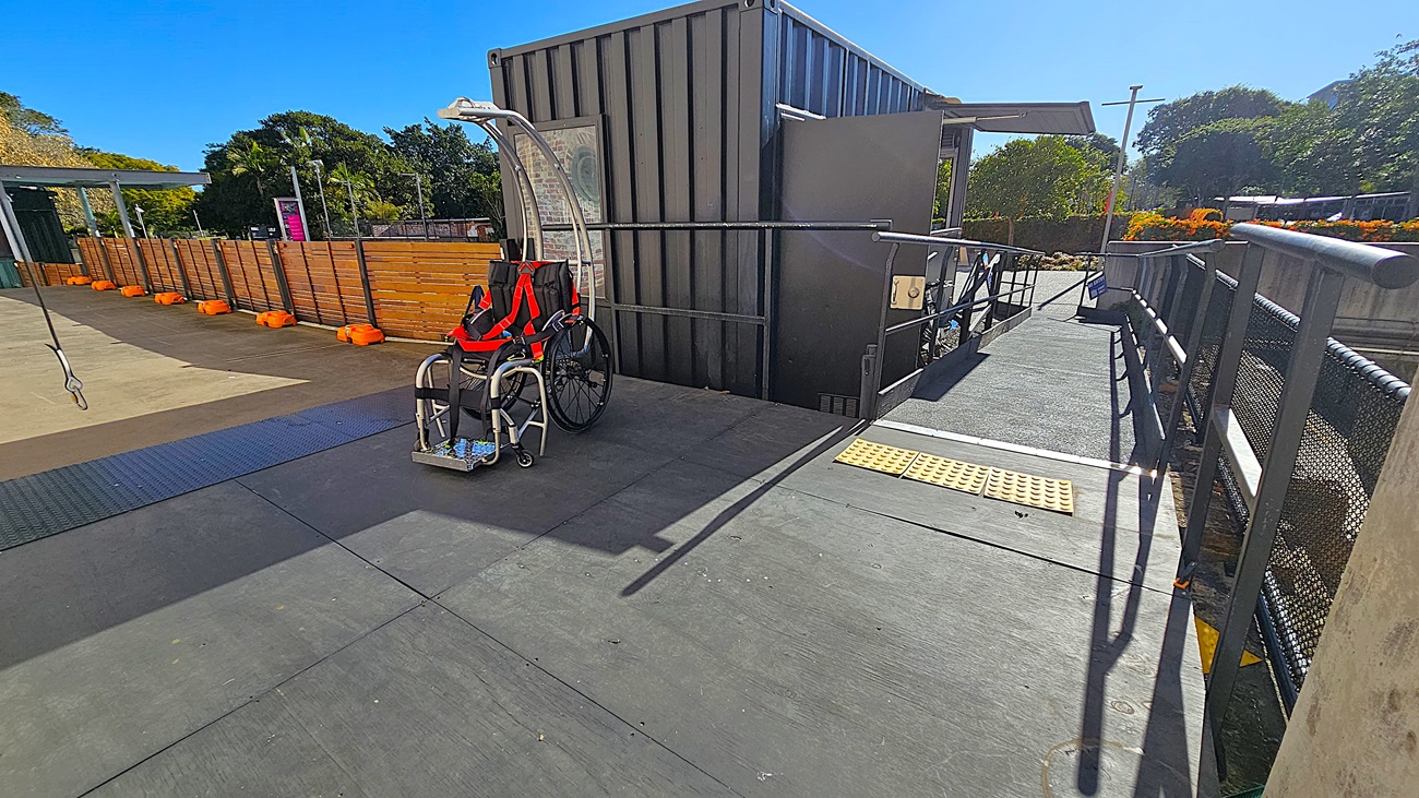 On the left, there is a black metal fence and a ramp leading up to a black structure with a sign reading "VERTIGO." Bicycles are parked next to the structure. In the middle, there is a blue carpet leading to the entrance of the structure. On the right, there are red metal tables and chairs on a textured concrete floor. In the background, there is a tall, weathered brick wall and trees. The floor is a mix of smooth and textured concrete.