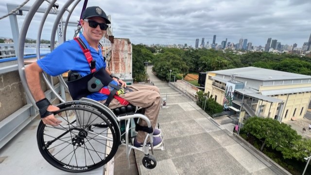 On the left, a man in a blue shirt and cap is seated in a wheelchair with a red harness, positioned on a metal platform with railings. In the middle, there is a wide concrete pathway leading to a large, weathered brick building. In the background, there are trees and a city skyline with tall buildings. On the right, there is a light-coloured building with a flat roof and multiple windows.