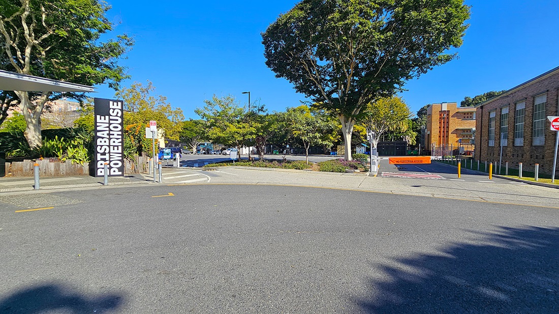 On the left, there is a sign reading "Brisbane Powerhouse" next to a building with a metal roof. In the middle, there is a paved road curving to the right, with a car park and trees in the background. On the right, there is a brick building with large windows and a "No Entry" sign. The floor is asphalt and concrete.