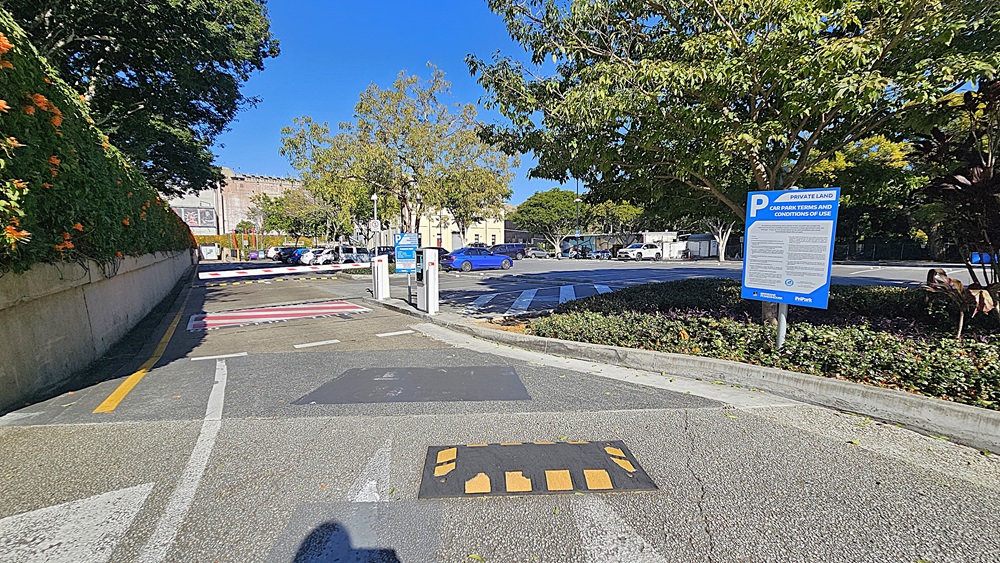 On the left, there is a concrete wall with greenery and orange flowers growing on it. In the middle, there is a paved road with a speed bump and a parking barrier, leading to a car park with several vehicles and trees in the background. On the right, there is a blue parking sign next to a landscaped area with bushes and trees. The floor is asphalt with painted markings.
