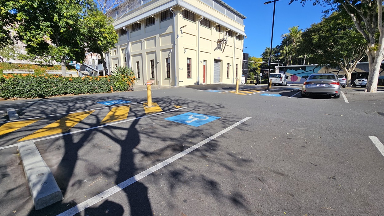 On the left, there is a row of bushes with orange flowers and a tree casting shadows on the ground. In the middle, there is a beige building with tall windows and a door, adjacent to a pedestrian crossing with yellow stripes. In the background, there are more trees and a mural on a distant wall. On the right, there are parked cars in marked bays, including a blue disabled parking space. The floor is asphalt with painted lines.