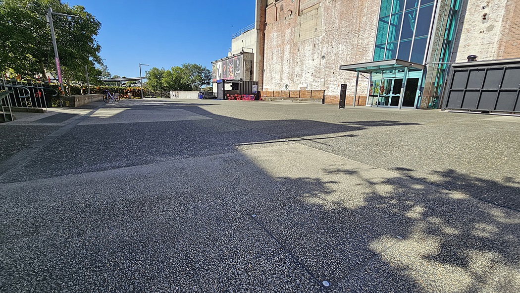 On the left, there are trees casting shadows over a textured pavement and a metal railing. On the right, there is a large brick building with tall windows and a glass entrance. In the middle, a wide, textured pavement leads towards the building. The background includes more trees, a small structure with a canopy, and part of the building. The floor is a textured pavement.